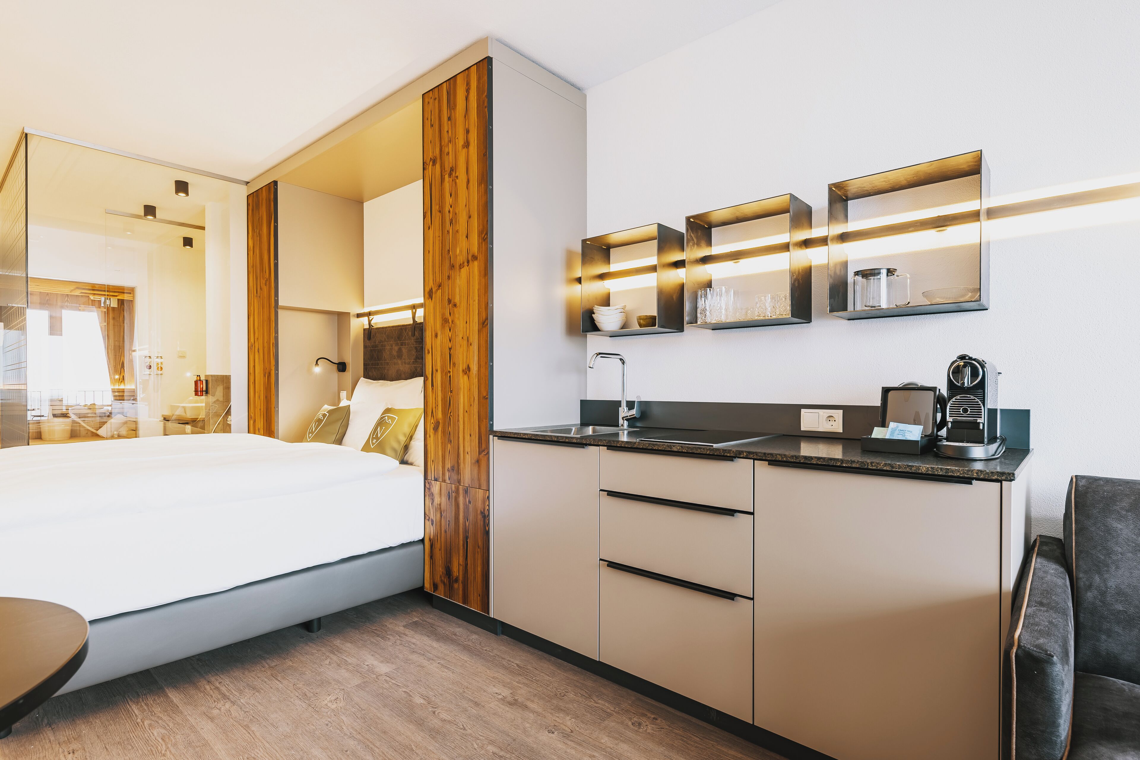 Una habitación de hotel moderna con una cama, una kitchenette pequeña y un baño visible a través de un separador de vidrio.