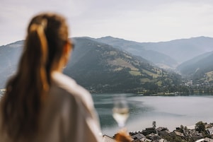 Alguém segura um copo de vinho apreciando a vista de um lago com montanhas ao fundo.