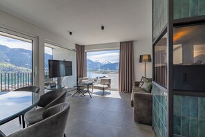 A modern hotel room with a large window, a flat-screen TV, a glass table, and a sofa. - Nikolaus by AvenidA Zell am See (Zell am See)