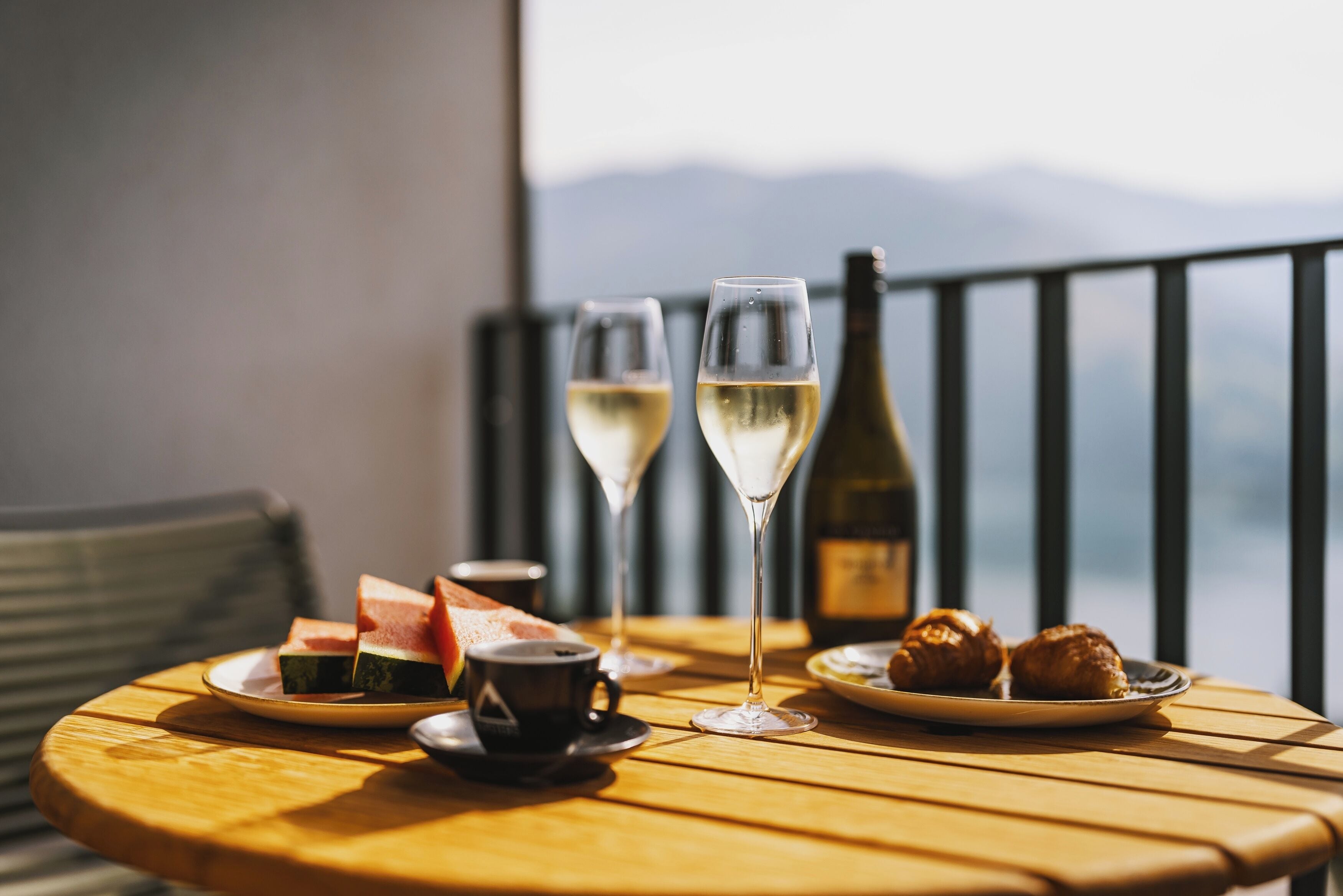 Una mesa de madera redonda con un plato de croissants, dos copas de vino blanco, una botella de vino, una taza de café y una rebanada de sandía.