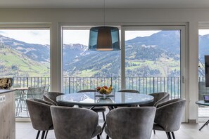 A round glass table with chairs, a fruit bowl, and a hanging light fixture in a room with a view of mountains. - Nikolaus by AvenidA Zell am See (Zell am See)