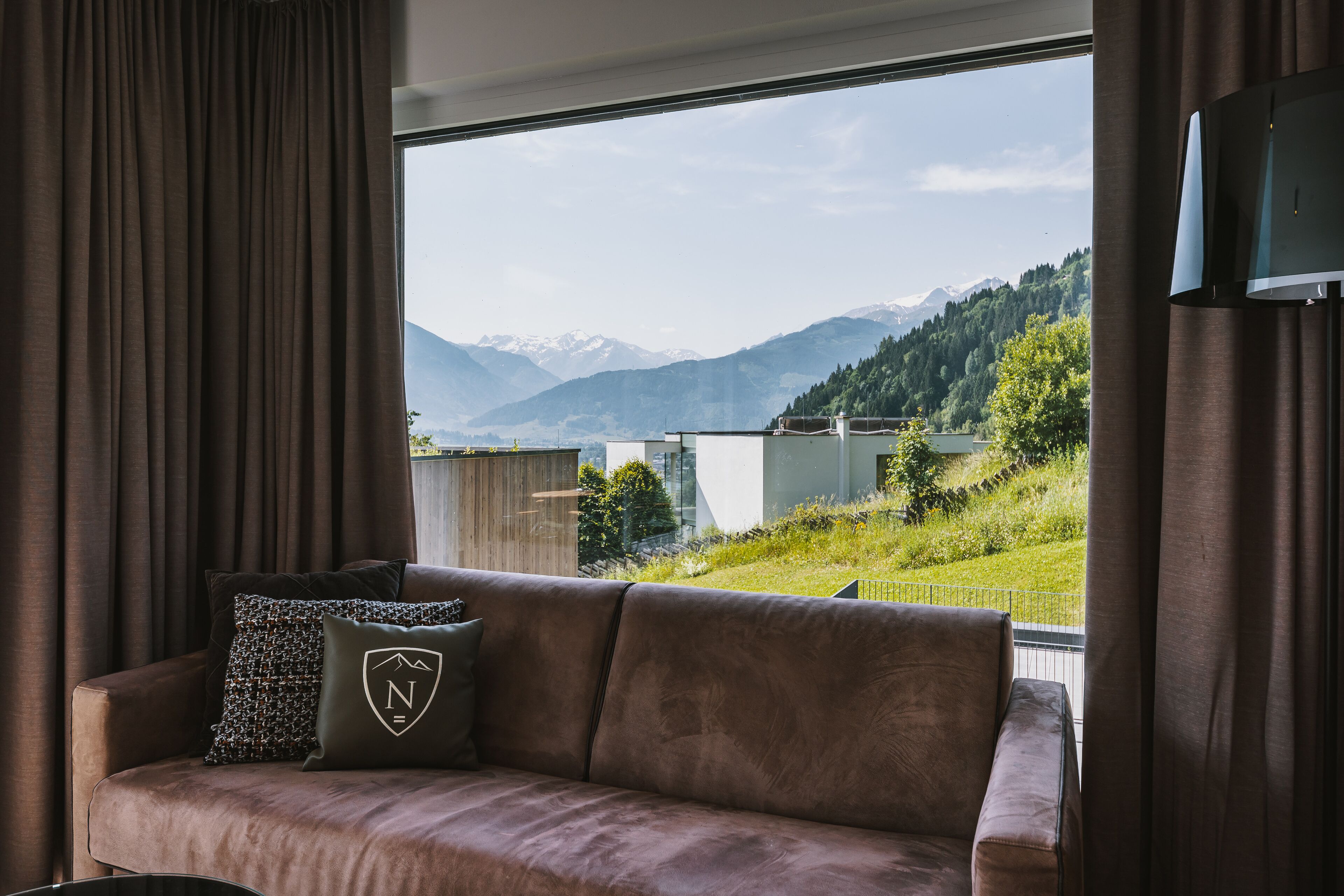 A room with a brown sofa, a window with a view of mountains and greenery, and a flat-screen TV mounted on the wall.