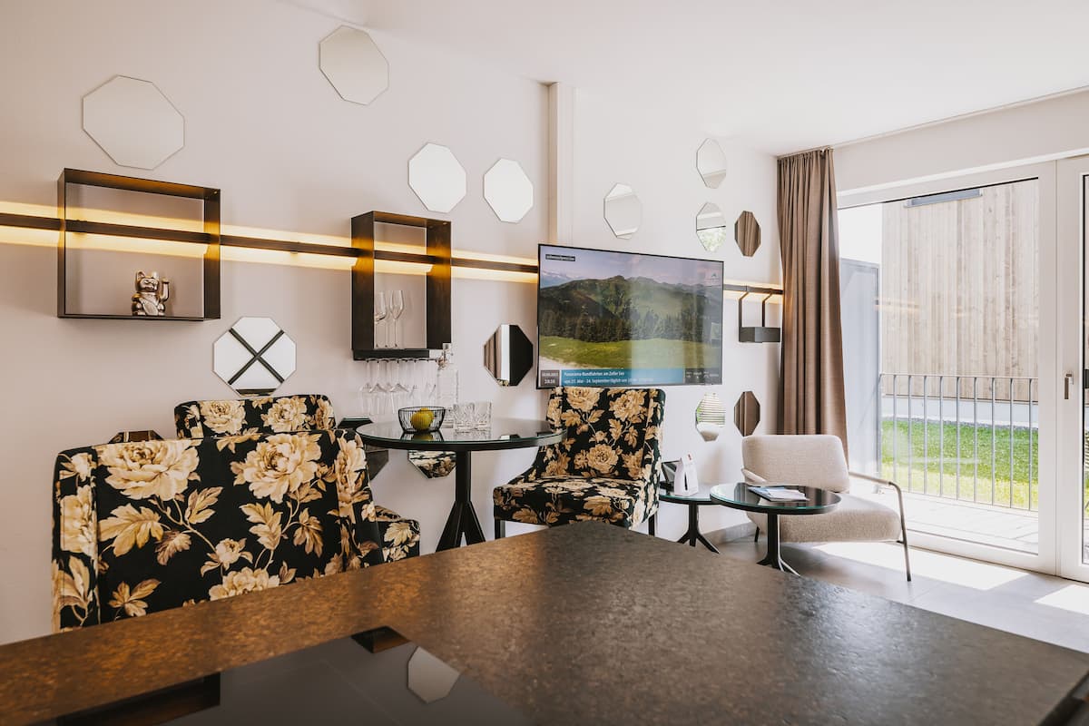 A modern hotel room with a large flat-screen TV, floral-patterned sofas, a dining table, and a balcony with a view of the outdoors.