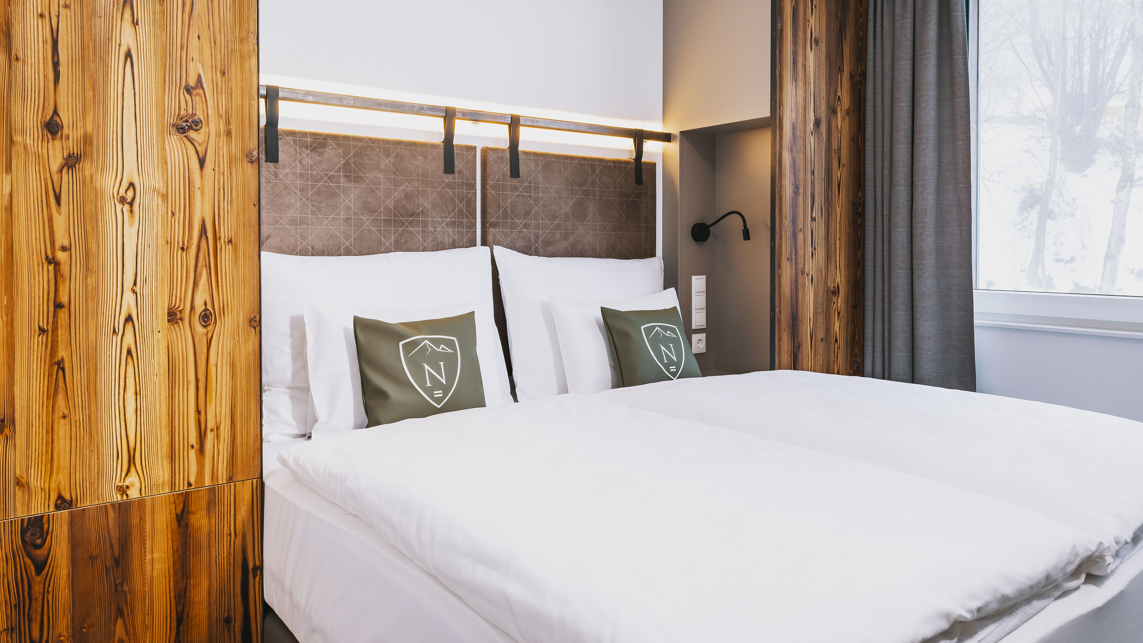 Una cama bien hecha con sábanas blancas, cabecera de madera y dos almohadas con el logo de una montaña.