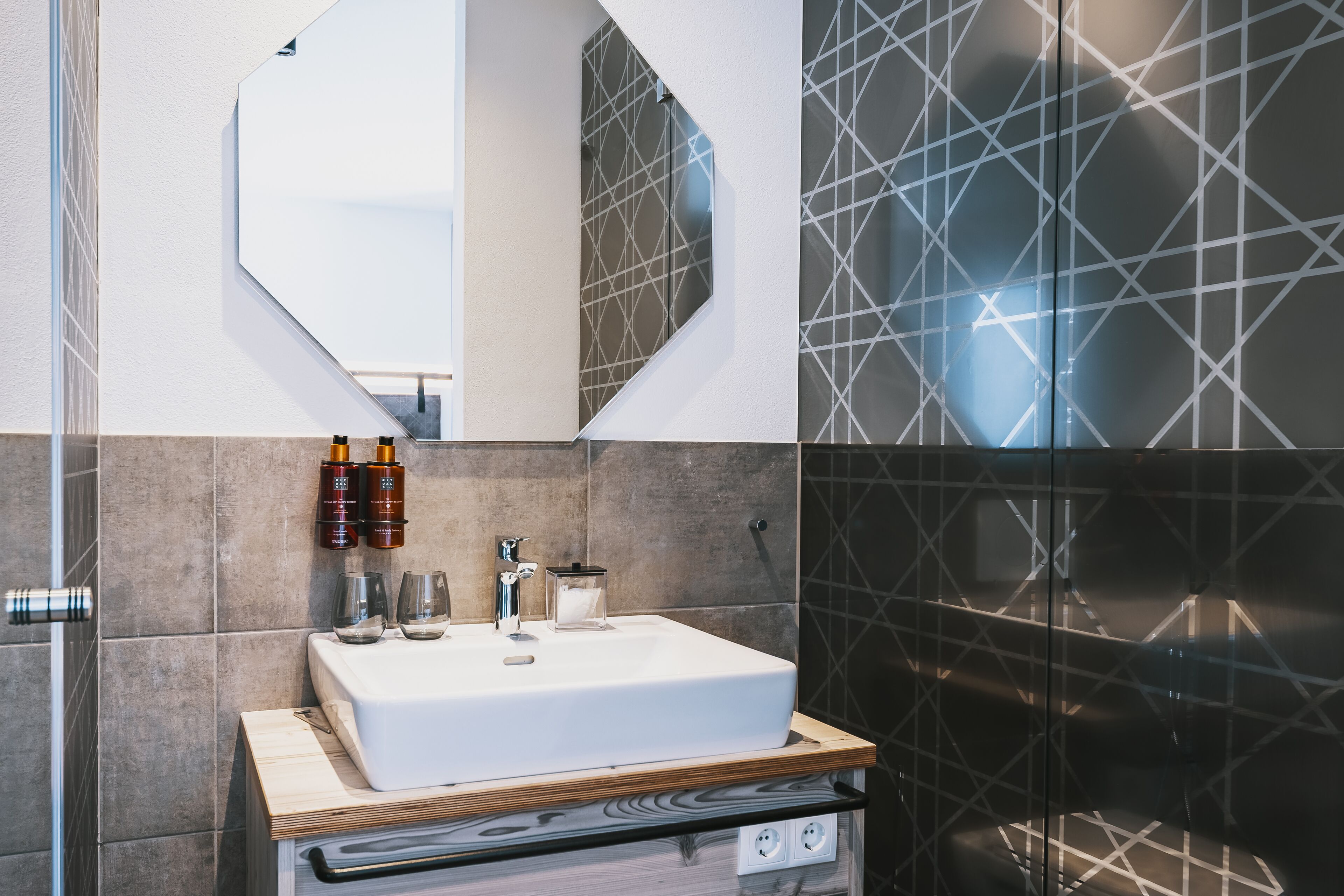A modern bathroom with a white sink, two mirrors, and a geometric-patterned wall.