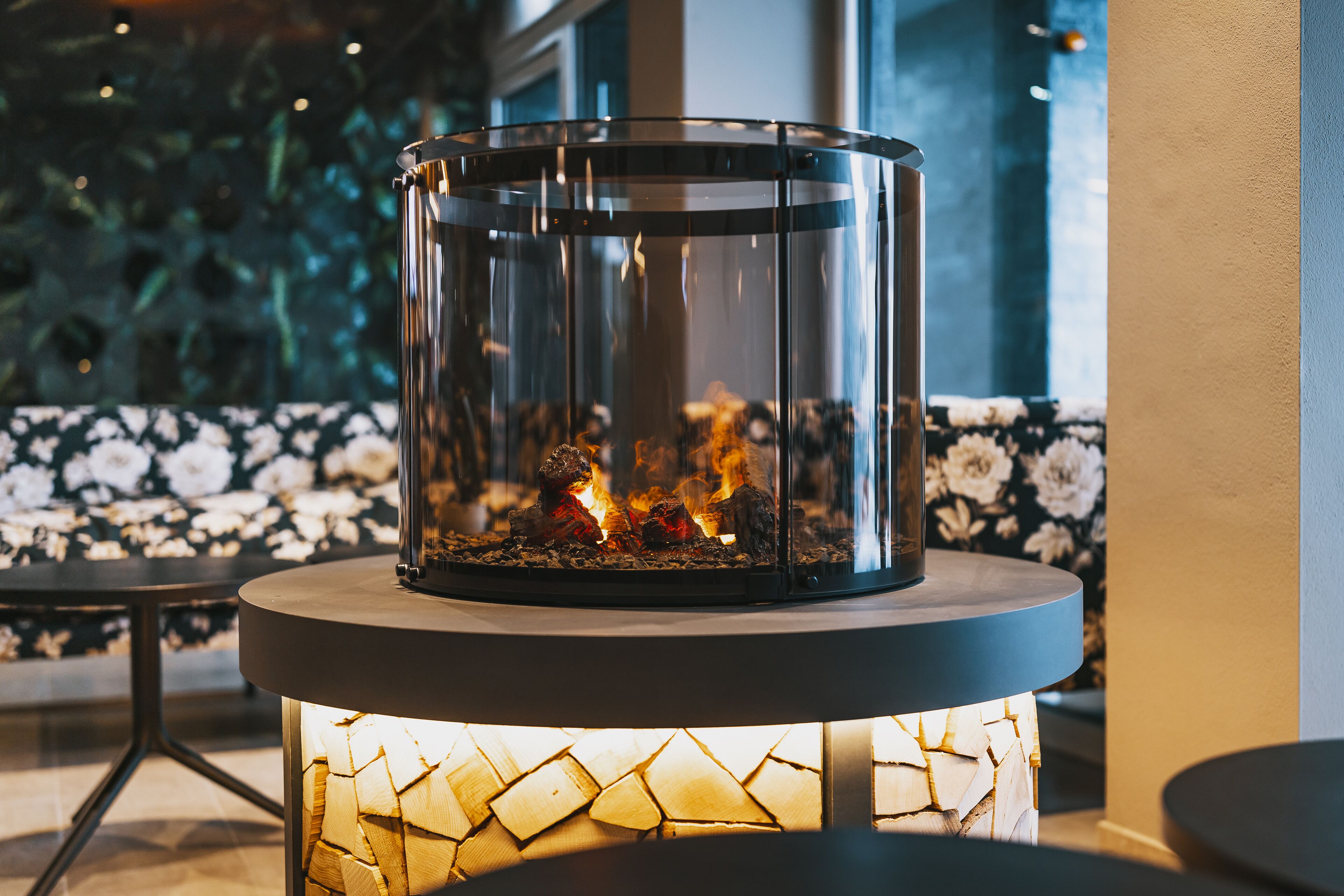 A glass-enclosed fireplace with visible logs and flames, set on a round table with a wooden base.