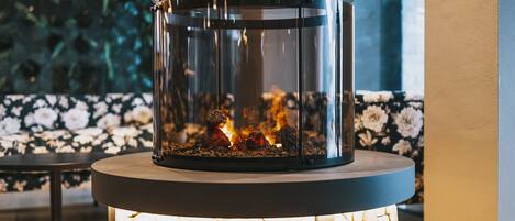 A glass-enclosed fireplace with visible logs and flames, set on a round table with a wooden base.