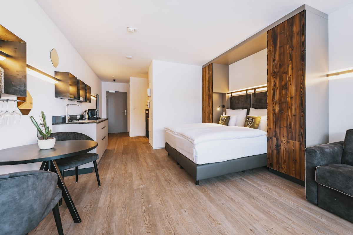 A modern hotel room with a bed, a desk, a chair, a small table with a potted plant, and a wooden accent wall.
