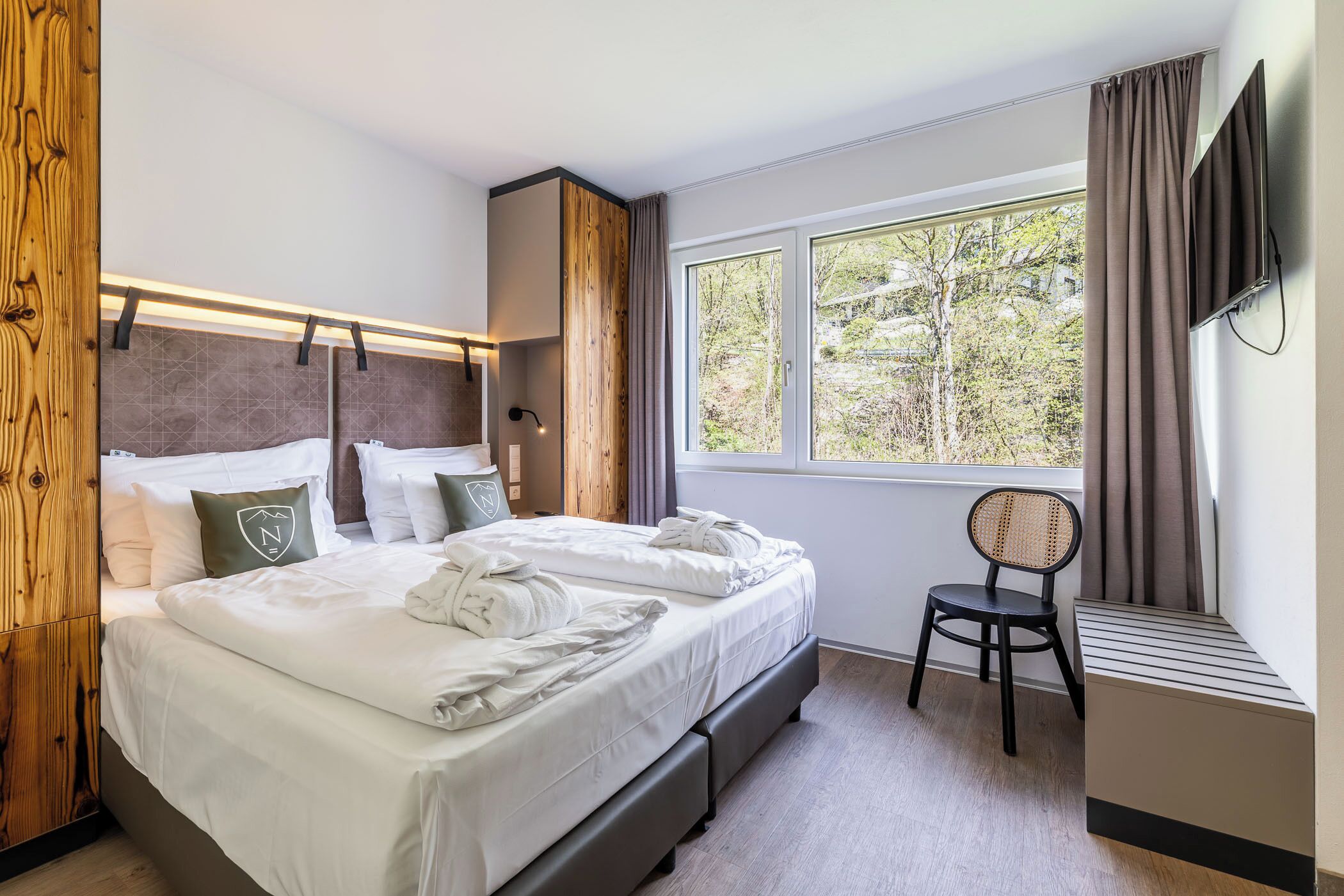 Ein Hotelzimmer mit Bett, Sessel, Fernseher und Fenster mit Blick auf Bäume.