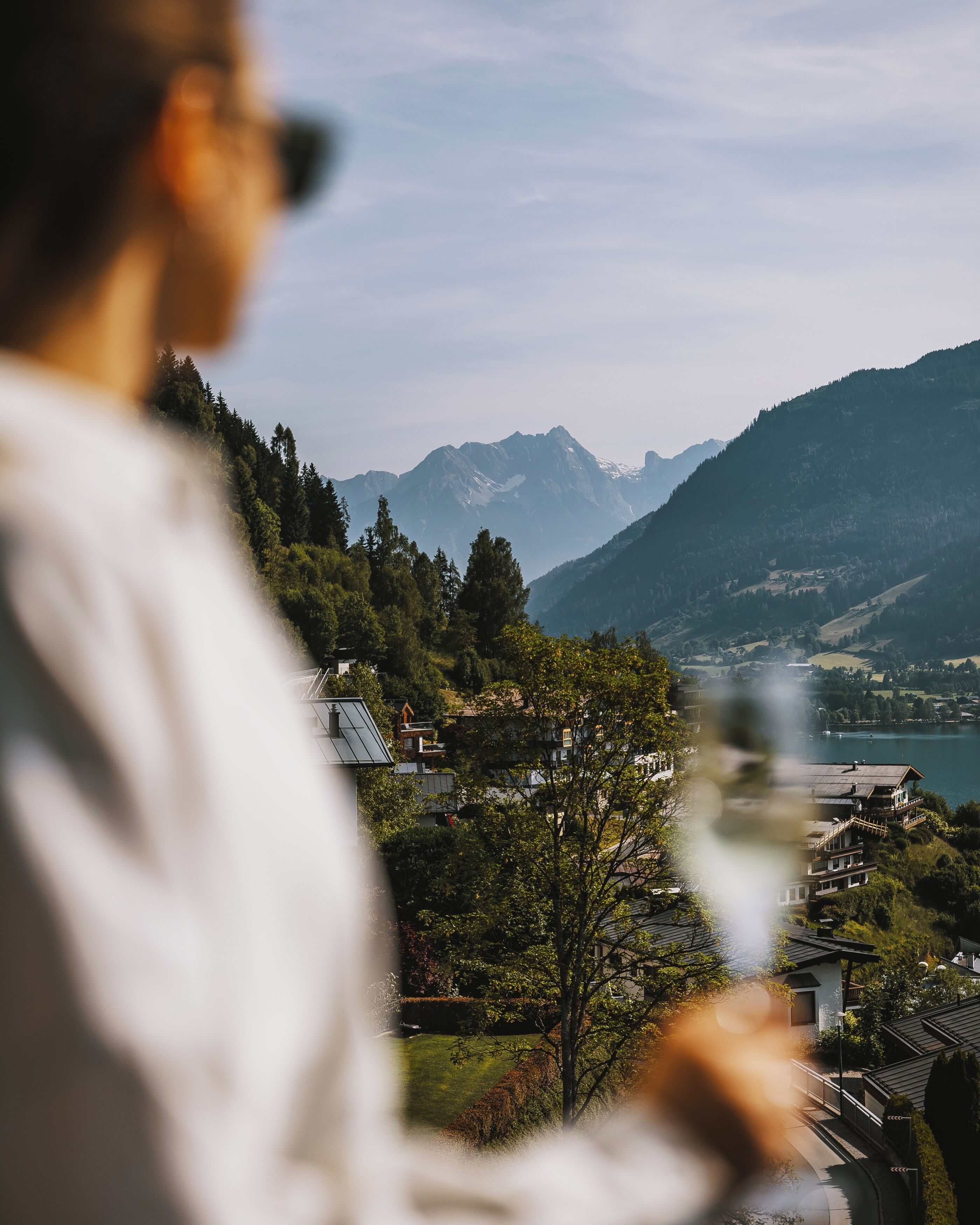 Una persona sosteniendo una cámara con vista a un paisaje montañoso, que incluye casas y un lago.