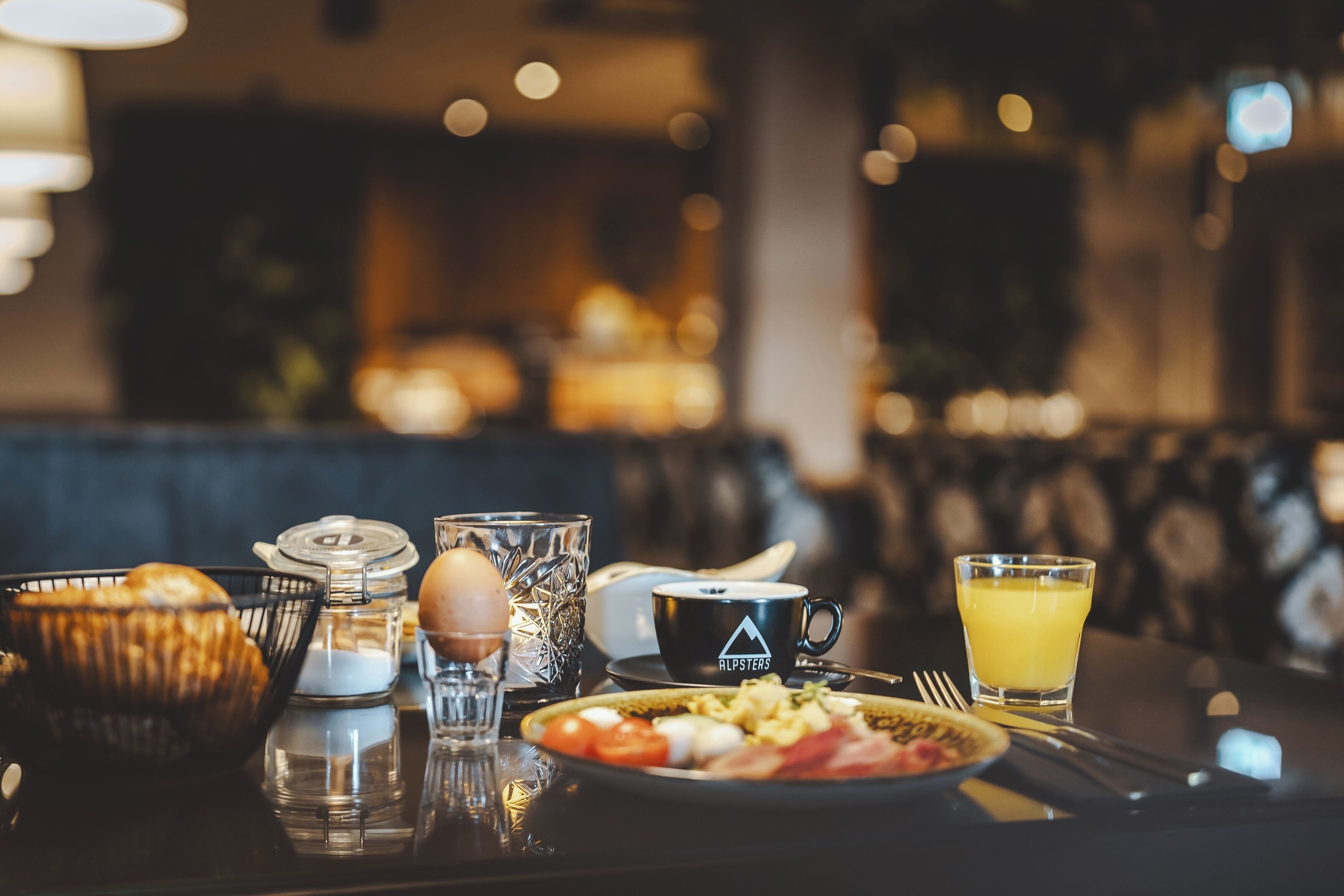 Et morgenbord med en tallerken mad, et glas appelsinjuice, en kaffekop, en kurv med brød og et glas vand på et bord.