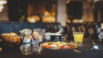 Et morgenbord med en tallerken mad, et glas appelsinjuice, en kaffekop, en kurv med brød og et glas vand på et bord.