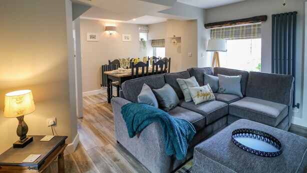 A living room with a grey sofa, a dining table with chairs, and a lamp on a side table. - The Stables (Stoke-on-Trent)