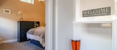 A bedroom with a bed, a nightstand, a lamp, and a 'WELCOME Please remove your shoes' sign.