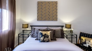Un lit bien fait, recouvert d’une couverture blanche à motifs, deux tables de chevet noires avec des lampes, un coussin décoratif et une œuvre d’art murale.
