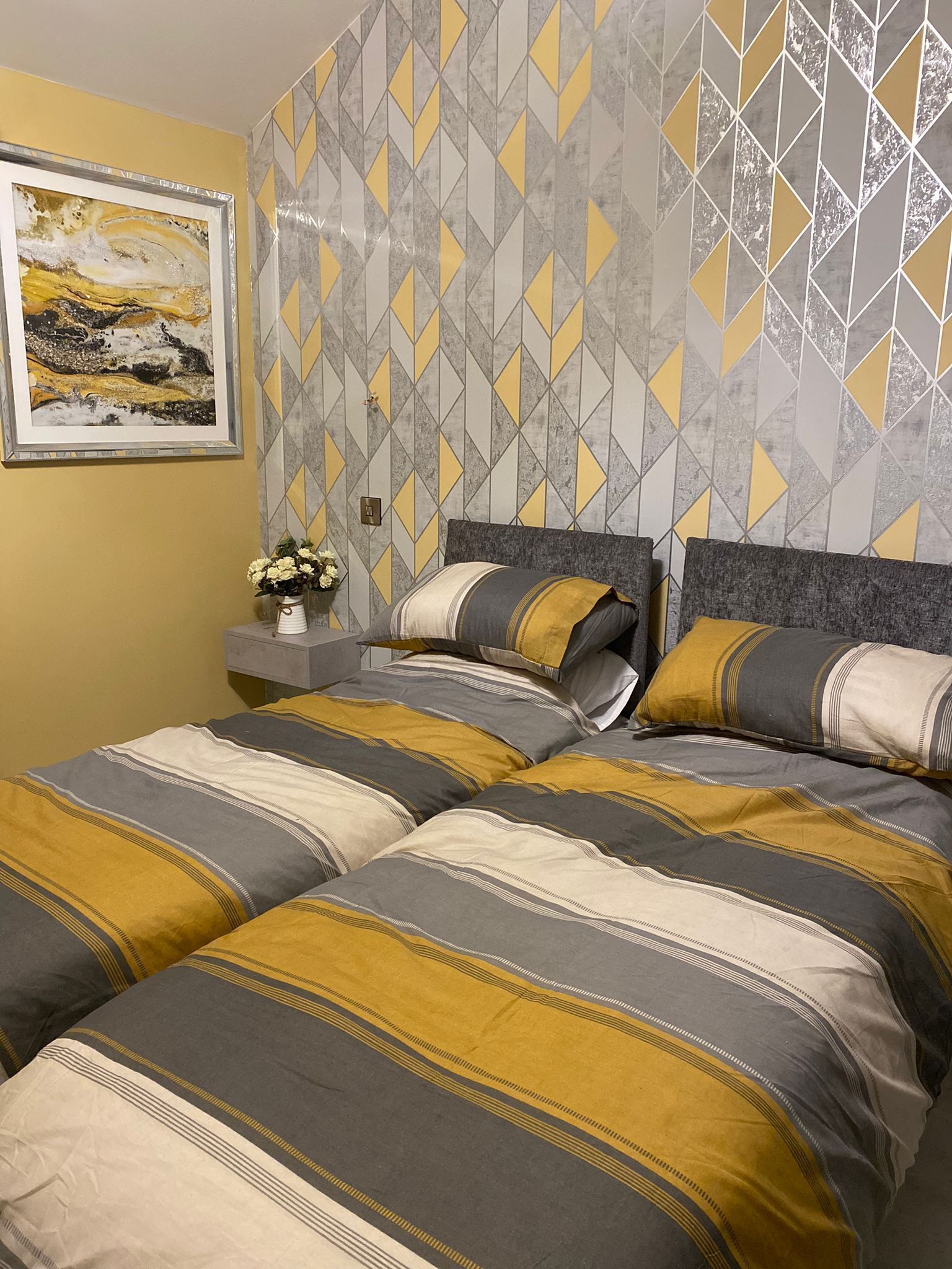 A bedroom with a geometric patterned wallpaper, a framed artwork on the wall, and a bedside table with a vase of flowers.