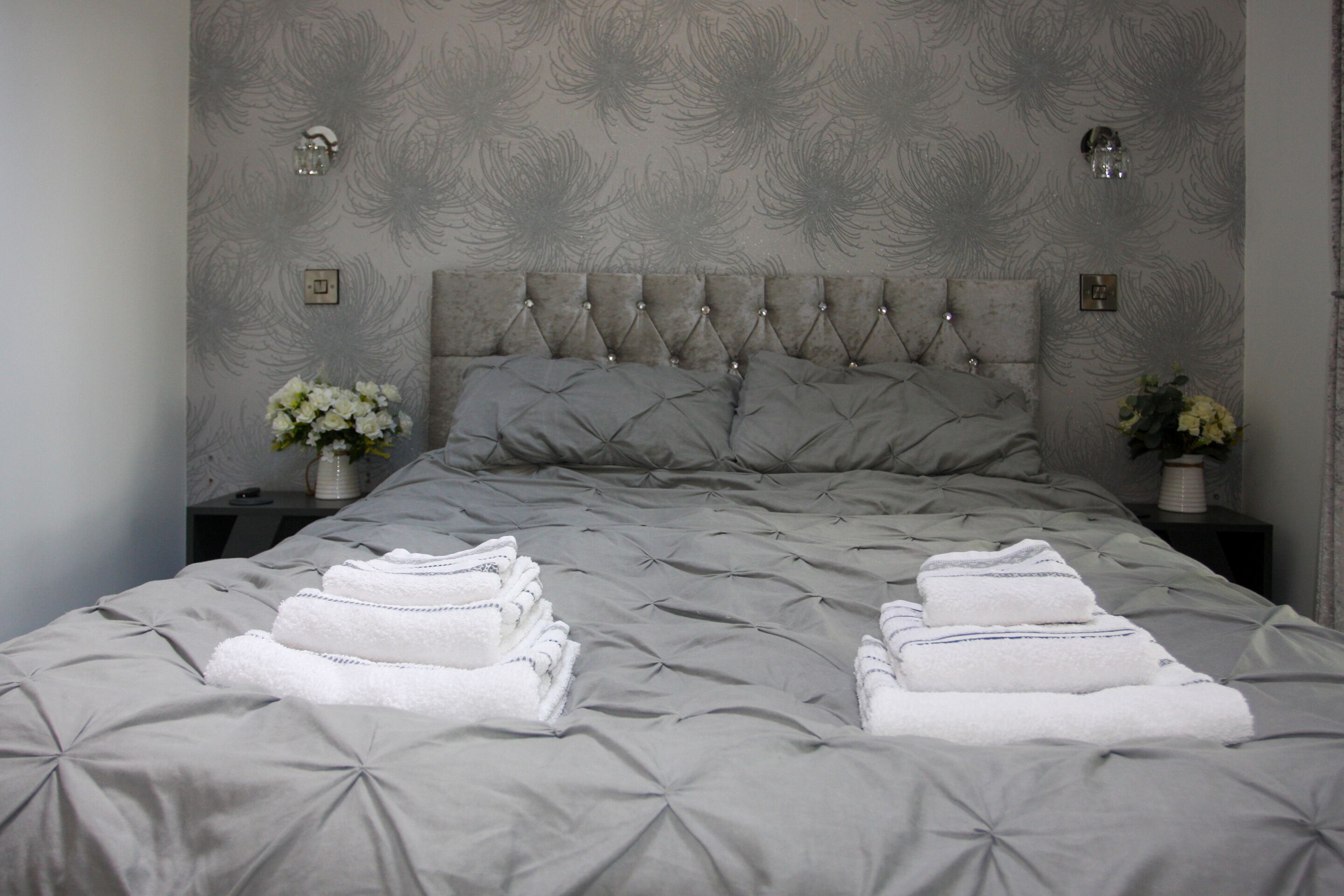 A neatly made bed with a grey quilt and pillows, two bedside tables with flower vases, and a wall with a floral pattern.