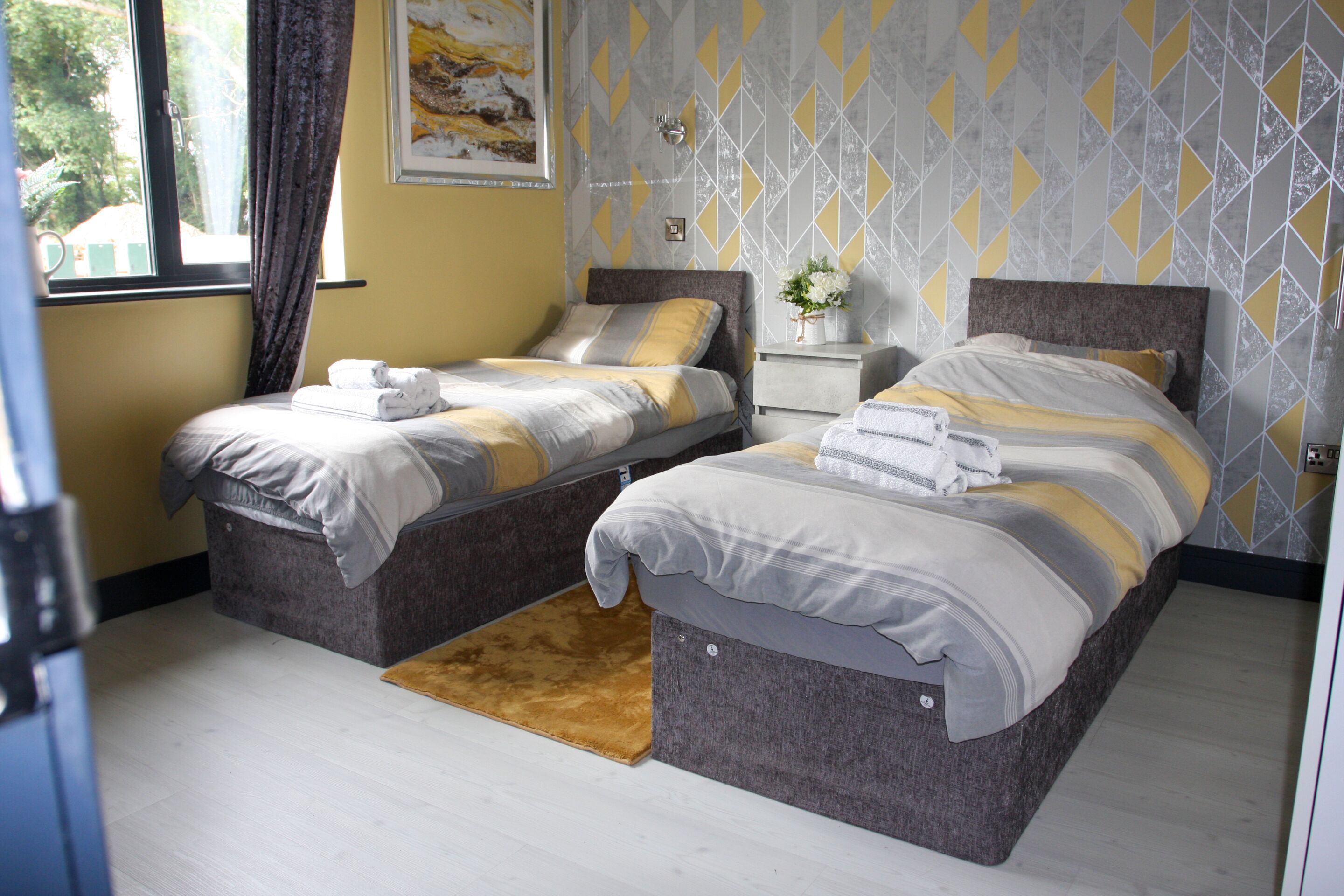 A hotel room with two beds, a small bedside table with a vase of flowers, and a window with curtains.