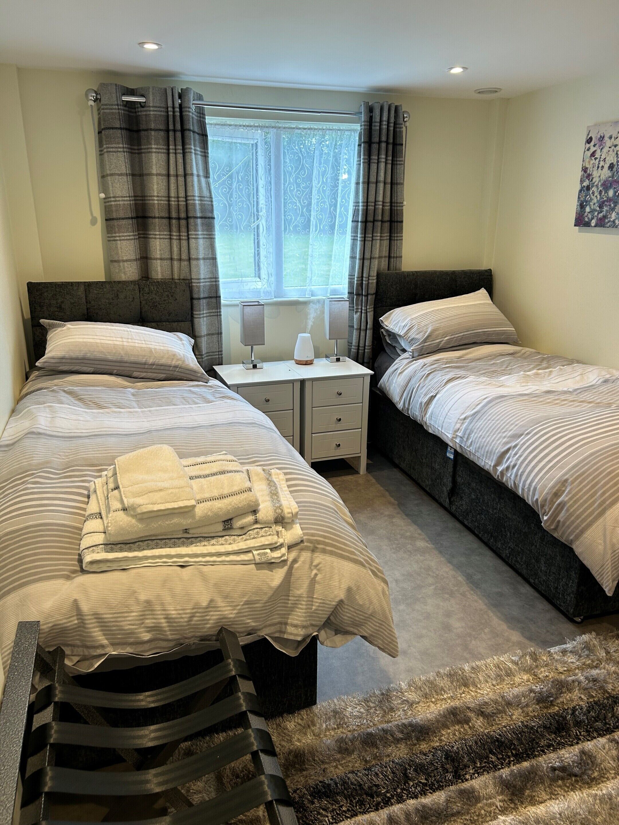 A hotel room with two beds, a window with curtains, a bedside table with lamps, and a small rug.