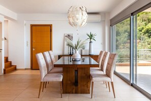 A modern dining area with a wooden table and cushioned chairs, a decorative light fixture, and a view of the outdoors through large windows. - Aigli Luxury Villa - Panoramic Seaview Retreat (Kalamata)