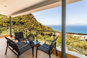 A balcony with a table and chairs, overlooking a coastal landscape with a view of the sea and a hill.