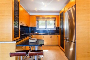 A compact kitchen with wooden cabinets, a stainless steel refrigerator, and a window with blinds.