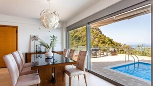 A modern dining area with a glass table and chairs, a decorative light fixture, and a view of a swimming pool and outdoor seating.