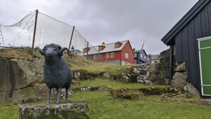 A stone sheep sculpture in a grassy area with a stone wall and a red building in the background.