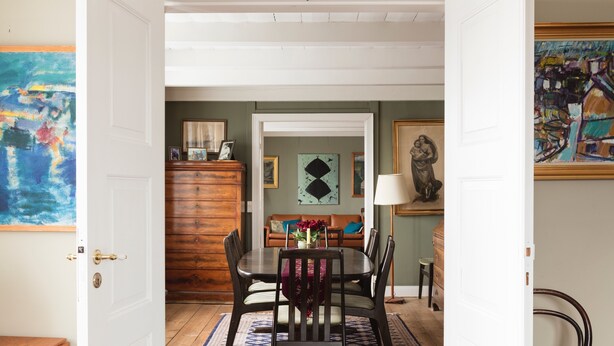 A dining area with a wooden table and chairs, a patterned rug, and a green wall with artwork.