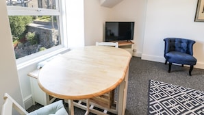 A room with a large window, a TV, a blue chair, and a patterned rug. - 4A Victoria Place (Barmouth)