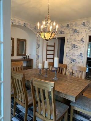 Un comedor con una mesa de madera, sillas, una araña y papel tapiz con un diseño azul y blanco.