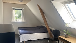 Ein Zimmer mit schräg gedachter Decke, einem Bett mit dunkelblauer Matratze, einem kleinen Tisch mit Topfpflanze und einem Fenster mit Vorhängen.