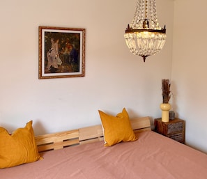 Un dormitorio con cama, dos almohadas, cabecera de madera, un cuadro enmarcado, una araña y una mesita de noche con lámpara y adorno.