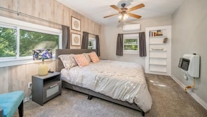 Ein Schlafzimmer mit einem Bett, einem Nachttisch, einer Lampe, einem Deckenventilator, einem Fenster mit Vorhängen, einem Bücherregal und einer Klimaanlage.
