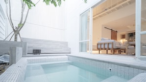 Piscina moderna al aire libre con plataforma azulejada, banca de madera y puerta de cristal que da a un espacio interior bien iluminado con sofá y mesa.