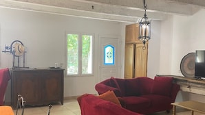 A spacious living room with a high ceiling, a wooden cabinet, a red sofa set, a dining table with a patterned tablecloth, and a television on a wooden stand. - Gite de charme 4 a 7 pers (Albaron)