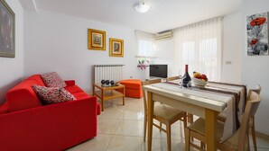 A modern living room with a red sofa, a dining table with chairs, and a kitchen area with a TV and air conditioning unit. - A one bedroom apartment with balcony and private parking in town - Dragica 2 (Umag)