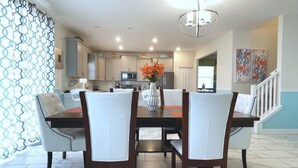 A dining area with a marble-tiled floor, a central table with chairs, a chandelier, and a kitchen in the background.