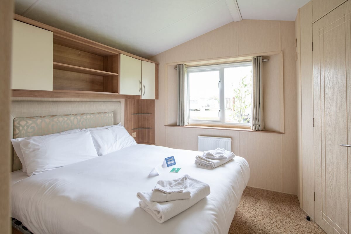 A hotel room with a bed, a window with curtains, a wooden wardrobe, and a small shelf above the bed.