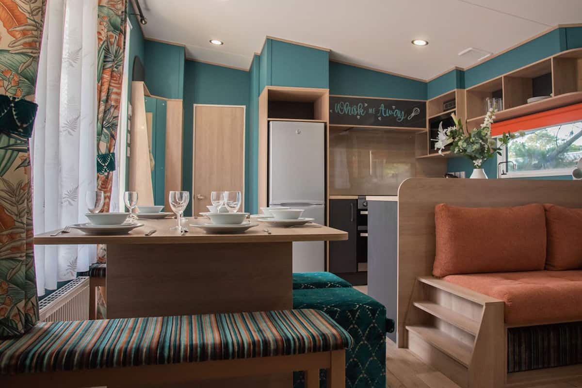 A compact, modern kitchenette with teal walls, a wooden dining table set with plates and glasses, and a cozy seating area with a cushioned bench and sofa.