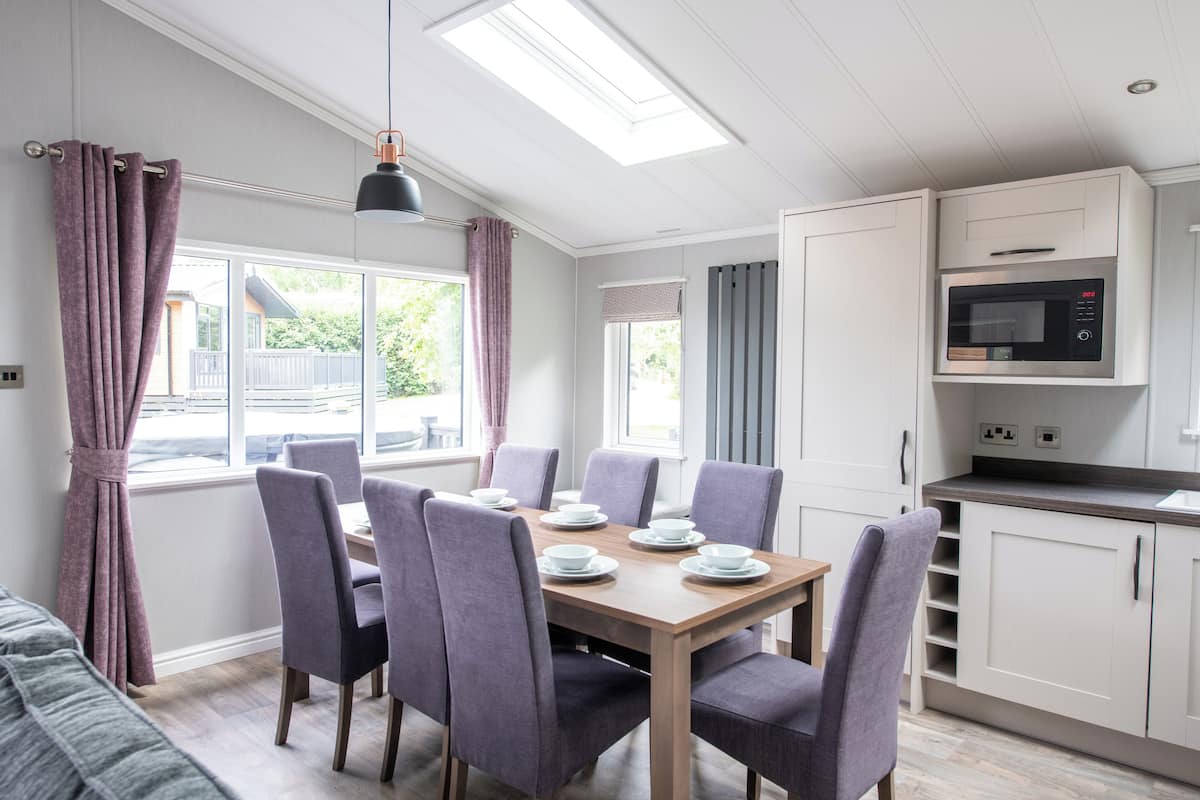 A dining area with a table set for six, six chairs, a ceiling light, and a kitchen with a microwave.