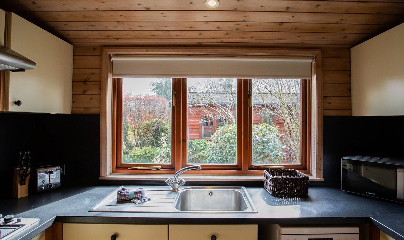 A kitchen with a large window, a sink, a microwave, and wooden cabinetry.