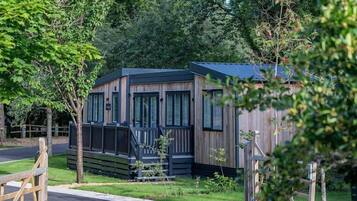 A small, modern cabin with a wooden exterior and large windows, surrounded by lush greenery.