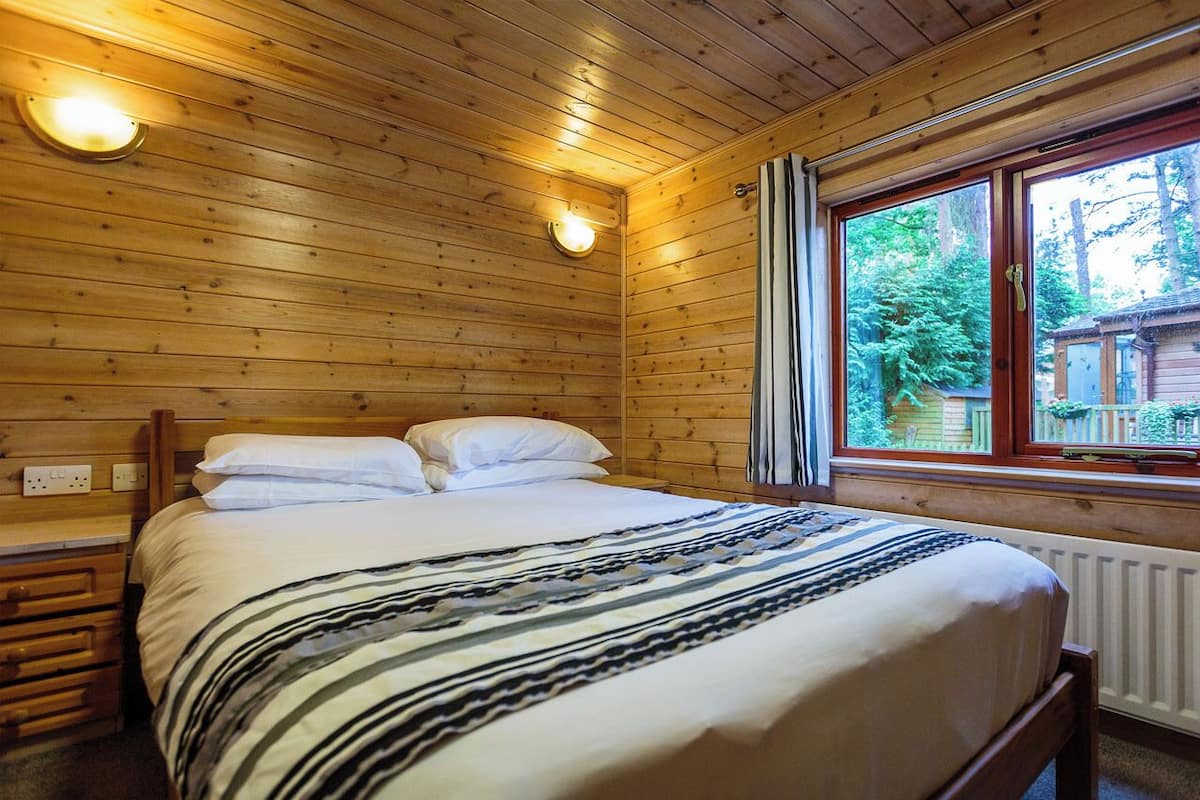 A wooden cabin room with a bed, two wall-mounted lights, a window with a view of trees, and a radiator.