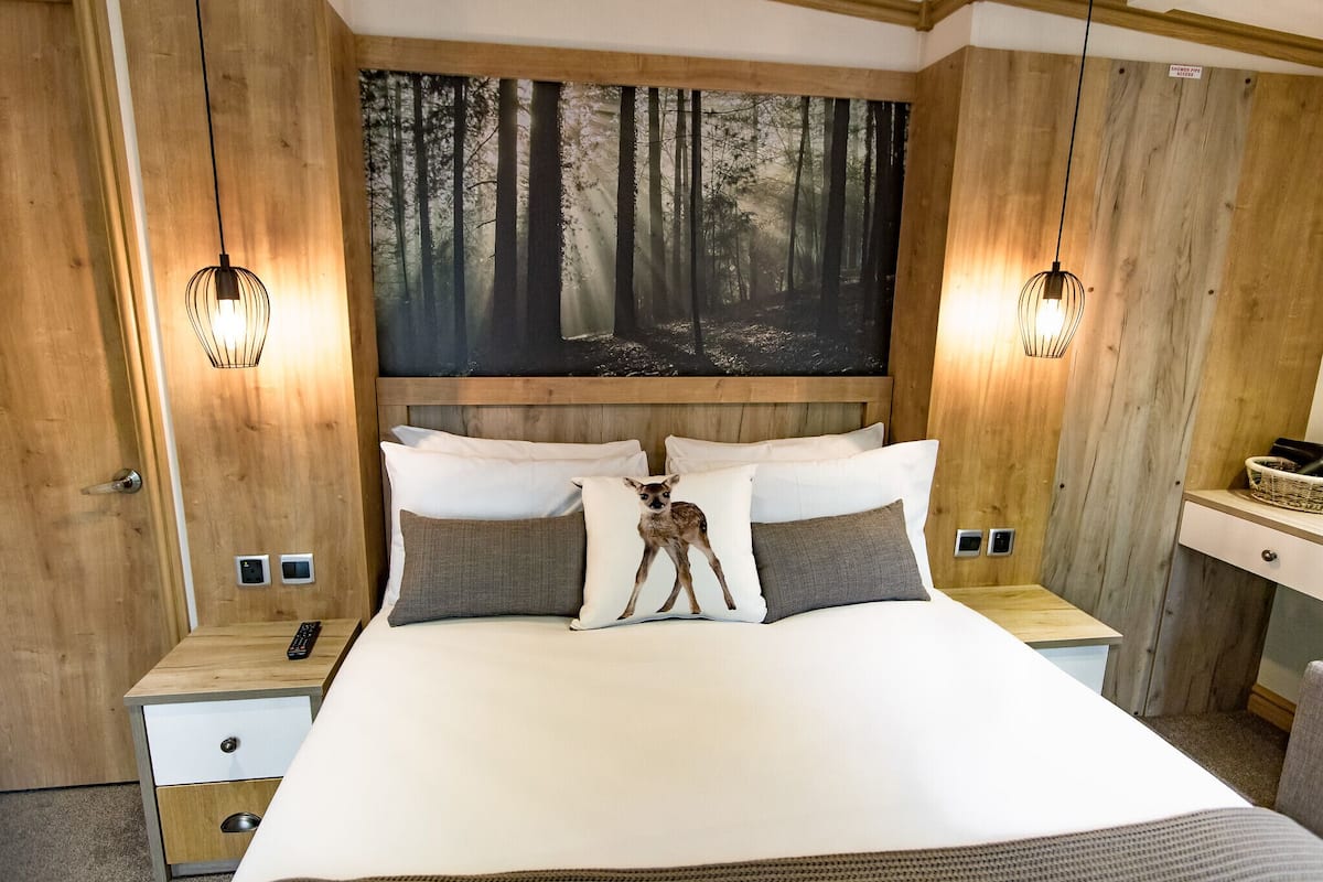 A bedroom with a large bed, two bedside tables, a nightstand, a wall-mounted painting of a forest, and two wall-mounted lights.