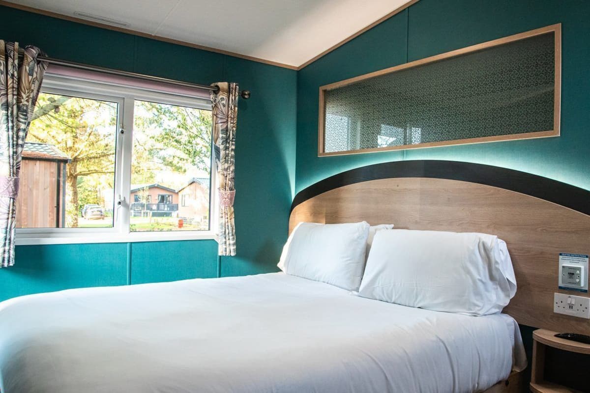 A bedroom with a large bed, a window with curtains, a bedside table, and a wall-mounted light fixture.