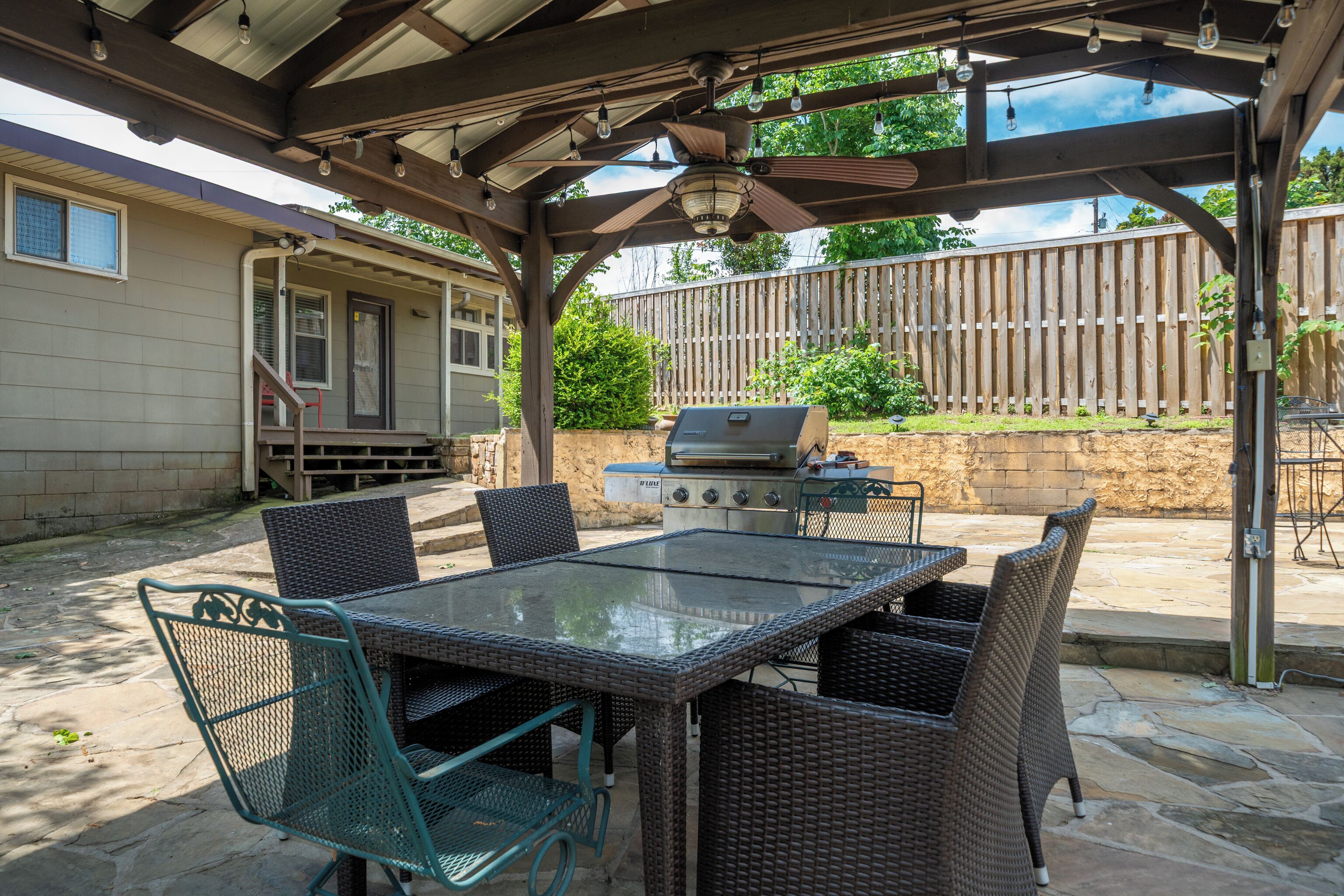 En uteplats med ett bord med glasskiva, flätade stolar, en grill och en pergola med belysning.
