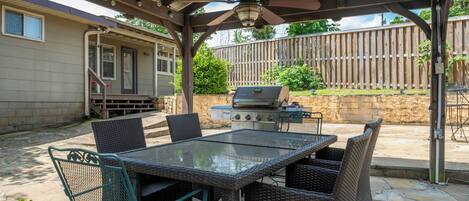 En uteplats med ett bord med glasskiva, flätade stolar, en grill och en pergola med belysning.