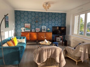 Una sala de estar con una pared azul estampada, un sofá azul, un mueble de madera, un televisor y una ventana con vista a follaje.