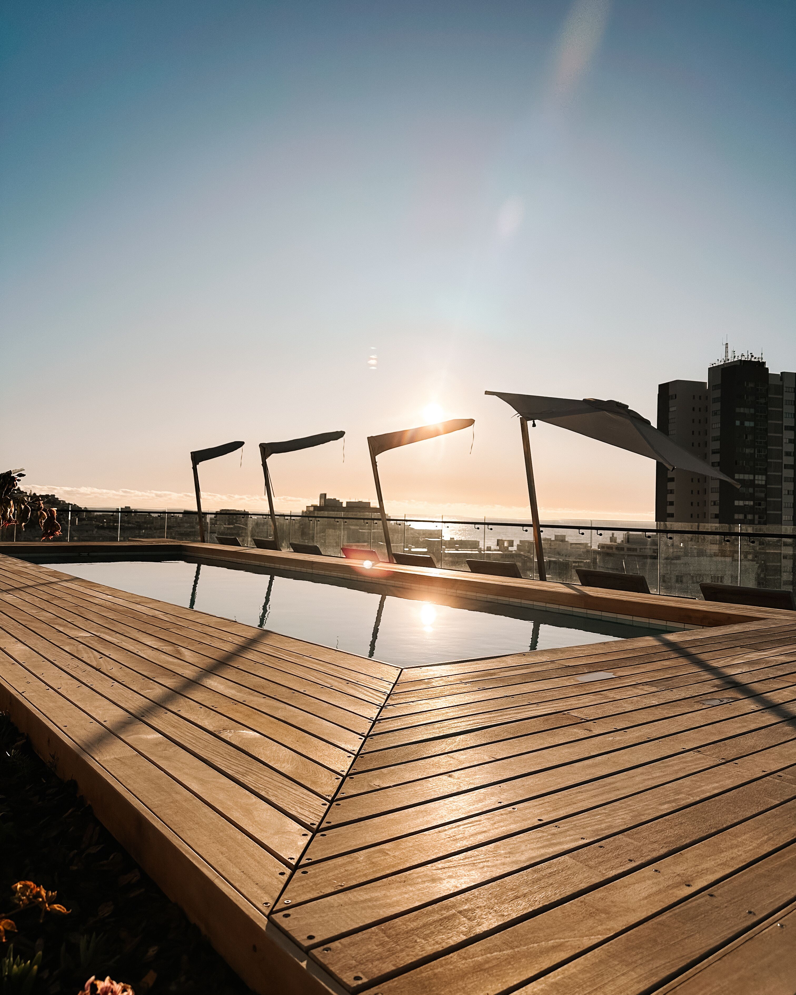 A rooftop pool with wooden decking and lounge chairs, overlooking a cityscape.